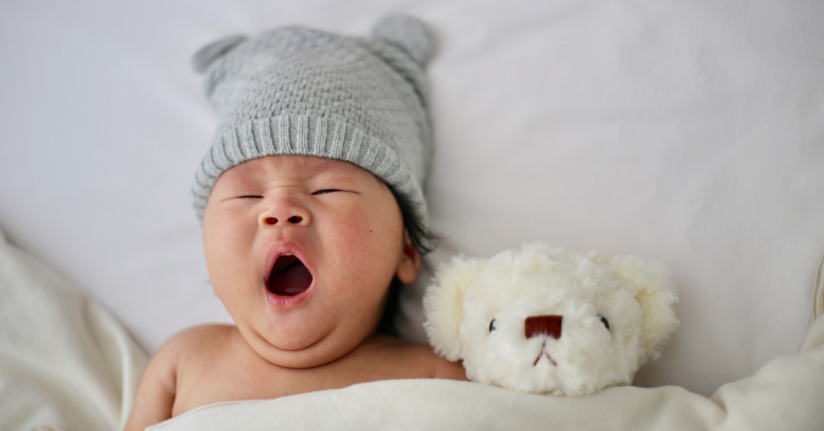 Baby with grey beanie yawning in bed next to teddy bear