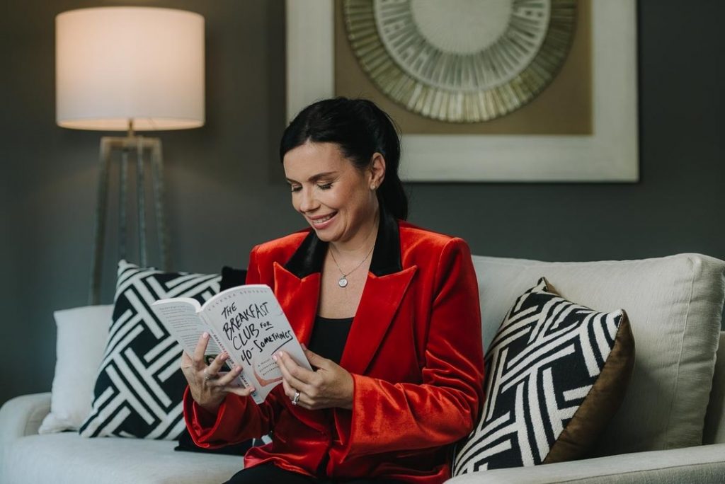 Vanessa Stoykov wearing a red jacket sits on a couch reading her book, The Breakfast Club for 40-Somethings,
